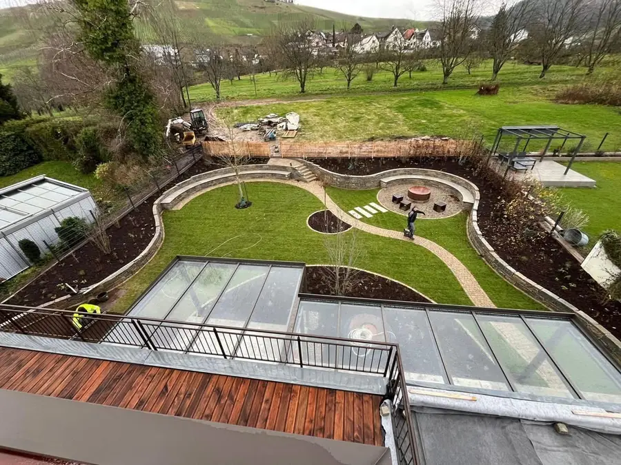 Blick von oben auf eine neu angelegte große Gartenanlage mit Natursteinmauern, geschwungenem Pflasterweg, Feuerstelle und Rollrase