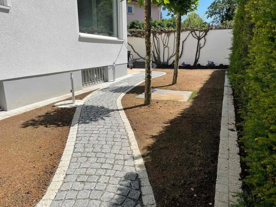 Frisch angelegter geschwungener Pflasterweg aus Kleinsteinpflaster mit neuem Beet, Mulchflächen und geschnittener Eibenhecke an ei