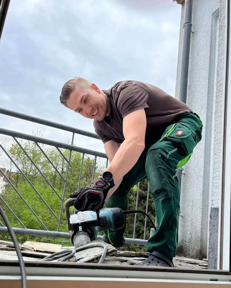 Lachender Mitarbeiter in Arbeitskleidung stemmt mit einem Makita-Abbruchhammer die alten Fliesen vom Balkon, Blick direkt in die K