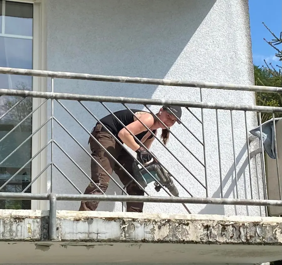 Mitarbeiterin mit Firmen-Cap stemmt mit dem Abbruchhammer den Balkonbelag ab, aufgenommen von unten durch das Balkongelaender. Auf