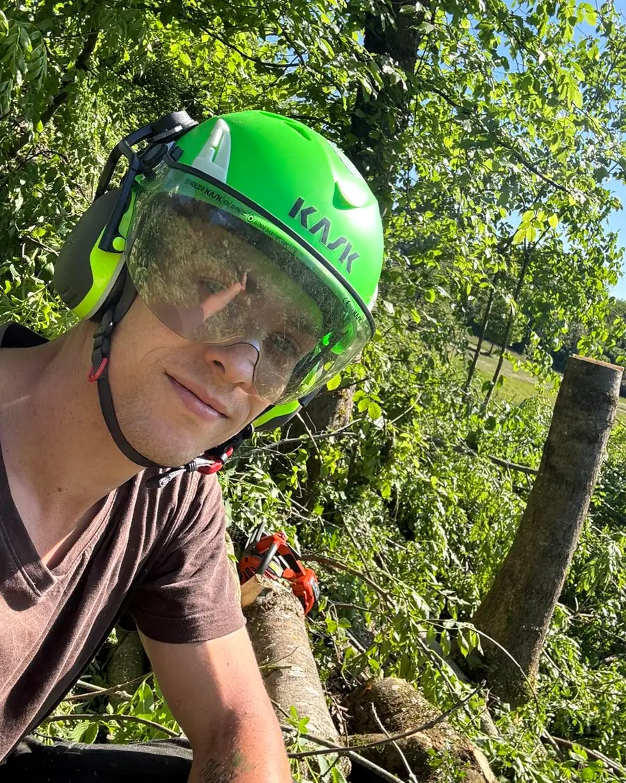 Selfie des Betreibers mit grünem KASK-Helm und Gehörschutz vor gefälltem Gehölz, im Hintergrund Motorsäge auf dem Stamm und frisch