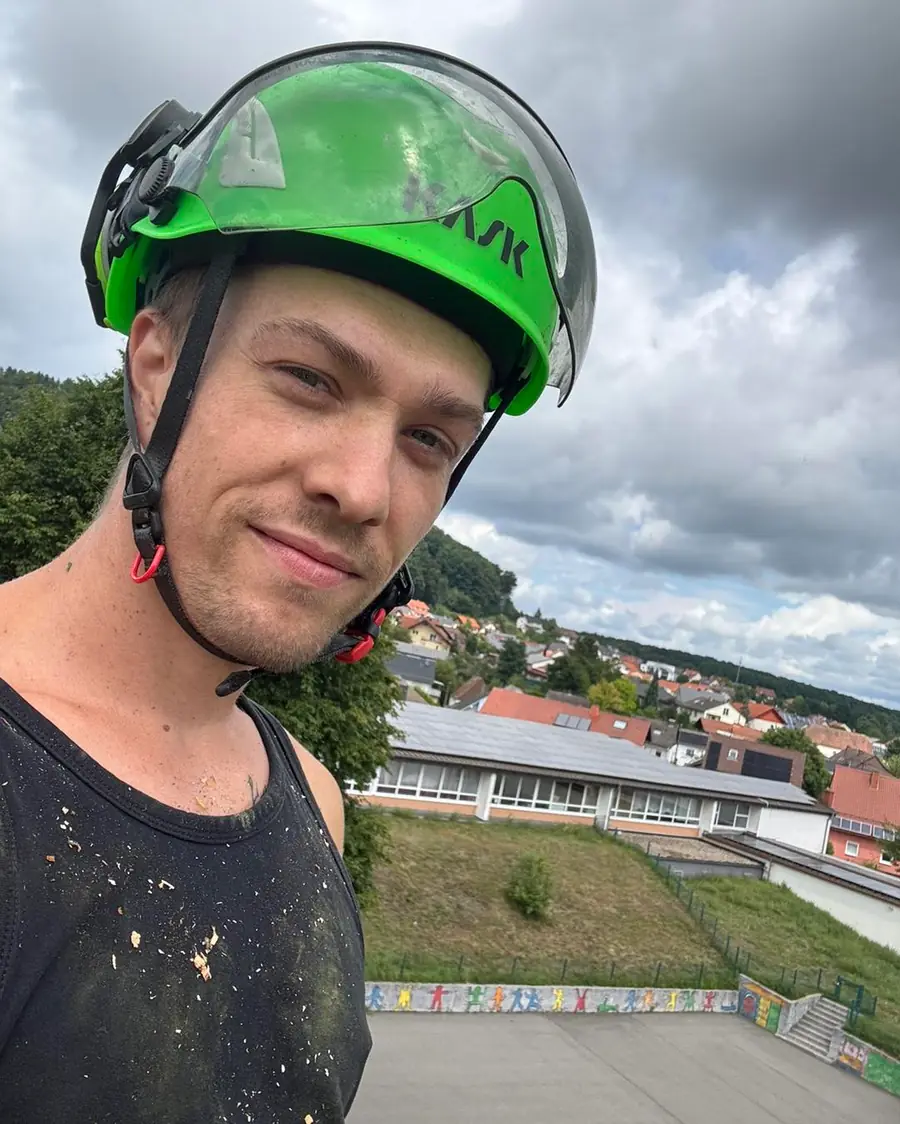 Selfie aus dem Baum mit grünem KASK-Kletterhelm, Sägespäne auf dem Shirt, weiter Blick über die Dächer des Ortes.